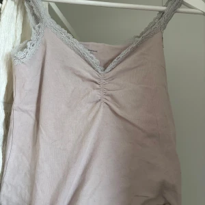 Ljusrosa linnetopp från Brandy Melville - Supersöt ljusrosa linnetopp från Brandy Melville med spetskant i grått och snygg rynkning vid v-ringningen. Mjuk bomullskänsla med lite stretch, perfekt för chill dagar. Axelbandslös look med romantiska detaljer som gör toppen extra gullig.