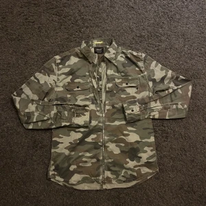 Camouflage overshirt från Replay - Säljer en snygg camouflage-overshirt från Replay med dragkedja framtill och två bröstfickor med knapp. Skjortan är i bomull och har klassisk krage och lång ärm. Perfekt för dig som gillar streetstyle och vill sticka ut med militärinspirerad look. Det står storlek L men jag skulle säga att den sitter mer som M.