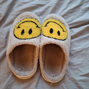 Grå tofflor med smiley-motiv - Mysiga grå tofflor med fluffigt tyg och stora gula smiley-ansikten broderade på ovansidan. De har en rund tå och platt sula i vitt, med mjuk insida för extra komfort. Perfekt för chill hemma och att glida runt i på morgonen.