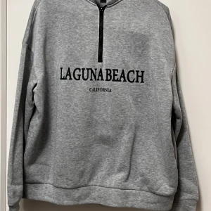 Grå sweatshirt med half zip från SHEIN - Säljer en grå sweatshirt från SHEIN i storlek XS med svart text 'Laguna Beach California' på bröstet. Tröjan har en half zip framtill och långa ärmar. Perfekt för chill dagar och enkel att matcha med jeans eller leggings.