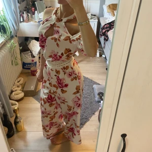 Vit byxdress med blommigt mönster - Säljer en vit byxdress med rosa och bruna blommor, snyggt axelbandslös med öppning vid bröstet och dekorativ spets i midjan. Byxdressen har ett luftigt fall och är perfekt för sommarens alla tillfällen. Materialet känns lätt och skönt mot huden.