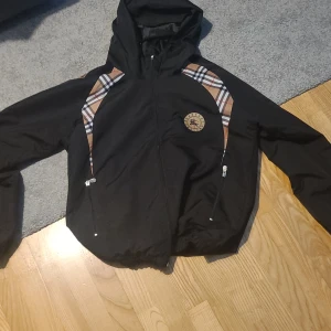 Svart vindjacka med rutig detalj - Svart vindjacka från Burberry med klassiska rutiga paneler på axlarna och insidan. Jackan har huva, dragkedja framtill och två fickor med dragkedja. Märkets logga på bröstet ger en exklusiv vibe. Perfekt för dig som gillar stilrena och tidlösa plagg.