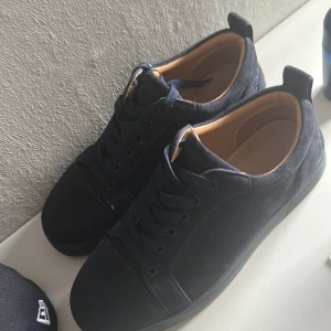 Mörkblå mocka sneakers från Louboutin - Säljer ett par stilrena mörkblå sneakers från Christian Louboutin i mjuk mocka med klassisk röd sula. Skorna har rund tå, snörning och diskret broderad logga på hälen. Insidan är beige och ger en lyxig känsla. Perfekt för dig som gillar exklusiva detaljer.