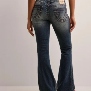 Blå bootcut jeans från True Religion - Säljer ett par snygga blå bootcut jeans från True Religion med klassiska kontrastsömmar och ikoniska bakfickor. Jeansen har låg midja och är tillverkade i stretchig denim för en bekväm passform. Perfekta för dig som gillar en retro vibe.