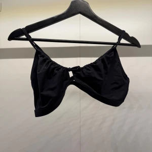 Svart bikiniöverdel med knyt från Ellos - Snygg svart bikiniöverdel från Ellos med justerbara axelband och knyt framtill. Modellen har en enkel och stilren design med metallspänne i ryggen. Materialet är mjukt och stretchigt syntet som sitter bekvämt. Perfekt för strandhäng och poolparty!