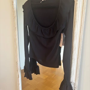 Svart blus med volang från Nelly - Supersnygg svart blus från Nelly med bred volang runt halsringningen och långa ärmar med volang vid ärmslutet. Helt ny med prislapp kvar