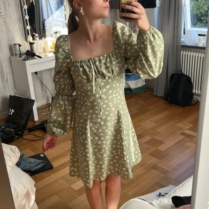 Grön blommig kortklänning med puffärm - Supersöt ljusgrön kortklänning med vita blommor, puffiga långärmar och fyrkantig urringning. Klänningen har snörning vid bysten och en diskret dragkedja i sidan. Materialet känns lätt och flowy, perfekt för vår och sommar. Passformen är normal och klänningen har en feminin siluett.