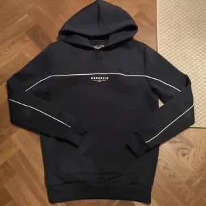  McKenzie hoodie - Snygg marinblå hoodie från McKenzie med vita linjer längs ärmarna och över bröstet. Tröjan har huva, ribbade muddar och ett stilrent tryck på bröstet. Perfekt för chill dagar och streetwear-stil. Materialet känns mjukt och skönt mot huden.