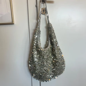 Silvrig axelväska med paljetter - Säljer en glittrig silvrig axelväska med massor av paljetter som täcker hela väskan. Den har en mjuk, rundad form och ett justerbart axelband i metallictyg. Perfekt för dig som vill sticka ut och addera lite extra bling till din outfit. 