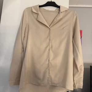 Beige långärmad skjorta med krage - Stilren beige skjorta med klassisk krage och knappar framtill. Skjortan har lång ärm och en rak, något längre passform baktill. Perfekt för dig som gillar enkla och cleana plagg. Materialet känns mjukt och lätt mot huden.