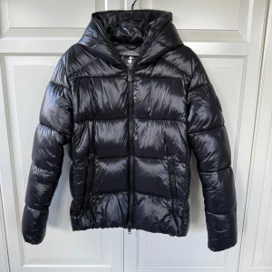 Svart pufferjacka från Save The Duck - Säljer en svart pufferjacka från Save The Duck med glansig finish och huva. Jackan är 100% animal free och har två dragkedjeförsedda fickor samt hel dragkedja framtill. Sparsamt använd, inga defekter och ser helt ny ut. Nypris 3699. 