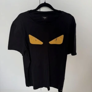 Svart Fendi t-shirt med gula ögon - Svart t-shirt från Fendi med ikoniska gula ögon framtill. Rund hals och korta ärmar. Mjuk bomullskvalitet och diskret Fendi-märke vid nederkanten. Perfekt för dig som gillar statement-plagg och designer.
