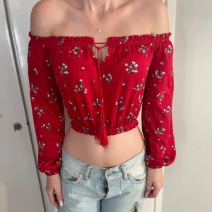 Röd blommig offshoulder croppad topp - Superfin röd croppad topp med blommigt mönster och offshoulder-design. Långa ärmar och smockad kant upptill och nedtill ger en snygg passform. Knytning framtill för extra detalj. Perfekt för dig som vill ha en söt och trendig look.