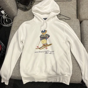 Vit hoodie Polo Bear Ralph Lauren - Vit hoodie från Polo Ralph Lauren med ikoniskt Polo Bear-tryck där björnen står på skidor i vinteroutfit. Tröjan har huva med snörning och långärmade muddar. Mjuk insida i bomullsmix, perfekt för chill dagar och cool stil.