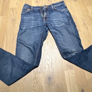 Blå Slim jeans från Nudie Jeans Co - Säljer ett par klassiska blå jeans från Nudie Jeans Co med snygg slitning och ett hål på ena benet. Jeansen har raka ben, fem fickor och orangea sömmar. Tillverkade i bomull med en cool tvättad look och läderpatch bak i midjan. I storleken W28 L32