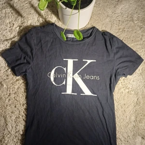 Calvin Klein Jeans T-shirt – Klassisk & stilren - Calvin Klein Jeans T-shirt – Klassisk & stilren  Säljer en snygg T-shirt från Calvin Klein Jeans. ✔️ Färg: Mörkblå / nästan svart med vit logga ✔️ Stor logga med CK Calvin Klein Jeans på bröstet ✔️ Material: Bomull (bekväm & mjuk) ✔️ Storlek: Längd-60cm & Axelbred-37cm ✔️ Skick: Mycket bra – inga hål eller fläckar, lite normalt tvättslitage  