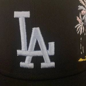 LA Dodgers svart keps 59FIFTY - Snygg svart LA Dodgers keps från New Era 59FIFTY med broderad LA-logga i ljusblått och palmer i pastellfärger på framsidan. MLB-logga broderad baktill. Klassisk formad skärm och stilren look, perfekt för dig som gillar streetwear.