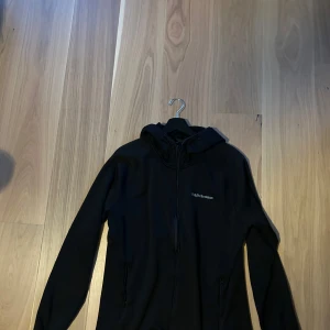 Svart hoodie/fleece från Peak Performance - Svart fleecehoodie från Peak Performance med dragkedja hela vägen och huva. Tröjan har lång ärm och två sidofickor med dragkedja. Snyggt broderad logga på bröstet. Perfekt för chill eller träning, stilren och enkel design.