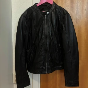 Svart skinnjacka med dragkedja - FUSK SKINN (vegansk) Snygg svart skinnjacka med raka linjer och cool ståkrage. Jackan har dragkedja framtill och två fickor med dragkedja på sidorna. Perfekt för dig som gillar en stilren och edgy look. Passar till jeans och boots för en klassisk vibe.