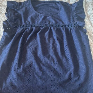 Mörkblå blus med volangärmar - Supersöt mörkblå blus med broderade detaljer och små hålmönster. Blusen har volangärmar och en rund halsringning. Ovanför bröstet finns en dekorativ blomspets som ger extra charm. Perfekt för dig som gillar romantiska och feminina vibes.