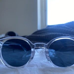Runda solglasögon med transparent båge - Snygga solglasögon med rund form och transparent plastbåge. Glasen är mörka och ger en cool kontrast mot den klara ramen. Perfekta för dig som vill sticka ut med en modern och stilren look. Passar till alla outfits och ger en trendig vibe.