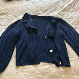 Mörkblå stickad kofta med volangkrage - Snygg mörkblå stickad kofta med volangdetalj vid kragen och pärlemorknappar framtill. Koftan har kabelstickat mönster på ärmarna och ribbade muddar. Perfekt för dig som gillar klassisk stil med en twist.