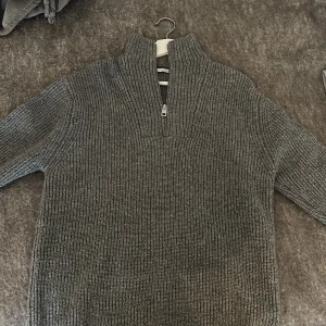 Grå stickad half zip tröja från Zara - Säljer en grå stickad tröja från Zara med ribbad struktur och hög krage. Tröjan har half zip-dragkedja framtill och långa ärmar, perfekt för kyliga dagar. Materialet är mjukt och värmande, och passformen är normal. Enkel att styla med jeans eller cargo.