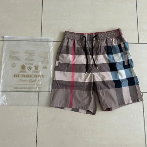 Burberry rutig shorts med snörning - Snygga Burberry shorts med klassiskt rutigt mönster i beige, brunt, svart, vitt och en röd detalj. Modellen har elastisk midja med svart snörning och praktiska fickor. Materialet är lätt och känns skönt mot huden, perfekt för varma dagar.