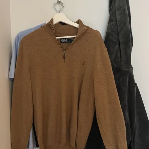 Brun stickad half zip från Polo Ralph Lauren - Säljer en brun stickad tröja från Polo Ralph Lauren med half zip och ribbad krage. Tröjan har långärmade ärmar och klassisk broderad logga på bröstet. Perfekt för dig som gillar stilrena och tidlösa plagg med snygg passform.lite små snoddar som åker ut men inte syns mycket