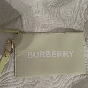 Ljusgrön kuvertväska från Burberry - Snygg ljusgrön kuvertväska från Burberry med vit logga framtill. Väskan har en avtagbar handledsrem med guldfärgad karbinhake och dragkedja upptill. Perfekt storlek för mobilen och det viktigaste. Stilren och modern design som lyfter din outfit.