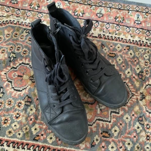 Svarta läder sneakers från XIT strl 39 - Svarta höga sneakers från XIT med snörning och dragkedja på sidan för extra smidig på- och avtagning. Mycket sköna skor för när man ska stå länge. Skorna har rund tå och platt sula, tillverkade i syntetislt läder med stilrena sömmar. Perfekt för dig som gillar en clean och urban look.