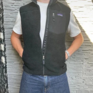 Svart fleeceväst från Patagonia - Svart fleeceväst från Patagonia med hög krage och hel dragkedja framtill. Västen har två sidofickor med dragkedja och en bröstficka med logga. Mjuk och varm, perfekt för lager-på-lager-stil under sommarkvällar eller kyliga dagar. Nypris 1400 kr, denna är så gott som ny! Fraktar oftast samma dag som köp😄