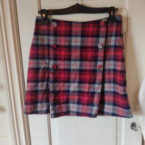 Rutig kortkjol med knappar - Säljer en rutig kortkjol i rött och blått med stora tygklädda knappar framtill. Kjolen har omlott-design och är fodrad med ett tunt mörkblått tyg på insidan. Perfekt för dig som gillar preppy och retrostil.