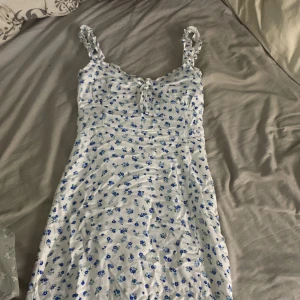 Vit blommig sommarklänning med volang - Supersöt vit klänning med blå blommor och volangdetaljer på axelbanden. Smockad rygg för skön passform och knyt framtill. Klänningen är kort och har en lätt, flowy känsla som passar perfekt till sommarkvällar, fester eller till vardag. Dragkedja på sidan och slits vis vänster ben. Använd 1 gång, pris kan diskuteras❤️