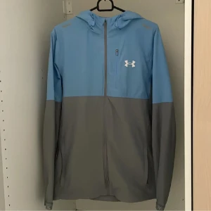Blå och grå vindjacka Under Armour - Snygg tvåfärgad vindjacka från Under Armour i blått och grått. Jackan har huva, hel dragkedja framtill och en liten bröstficka med zip. Perfekt för dig som gillar sportig stil och vill ha något lätt och funktionellt.