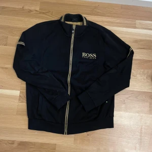 Svart ziptröja från Hugo Boss - Svart ziptröja från Hugo Boss med guldfärgade detaljer och logga på bröstet.Storlek M men passar S, inga defekter fint skick.