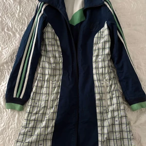 Adidas lång vindjacka med rutor - Snygg lång vindjacka från Adidas med marinblå bas och rutiga paneler i vitt, grönt och blått. Jackan har klassiska Adidas-ränder på ärmarna, gröna muddar och dragkedja framtill. Perfekt för dig som gillar sportig och retro stil. Tvåsidig vindjacka. 