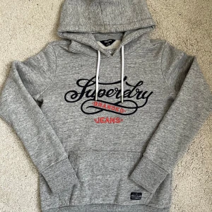 Superdry hoodie - Snygg grå hoodie från Superdry med stort svart och rött tryck på bröstet. Tröjan har huva med vita snören, ribbade muddar och en klassisk känguruficka framtill. Perfekt för chill dagar och streetstyle. Mjuk insida och skön passform. Passar som XS.
