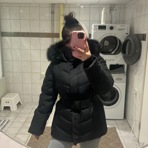 Svart pufferjacka med fuskpäls från Amisu - Säljer en svart pufferjacka från Amisu med stor huva och svart fuskpäls. Jackan har bälte i midjan, dragkedja och knappar framtill samt långa ärmar. Perfekt för kalla dagar och riktigt snygg siluett. Man kan även ta bort bältet. Helt oanvänd jacka!! 