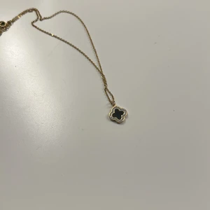 Guldhalsband med svart fyrklöver - Supersnyggt halsband i guld med en liten fyrklöverformad berlock i svart. Runt berlocken sitter glittriga stenar som ger extra bling. Kedjan är tunn och elegant, perfekt för att lyfta din outfit med en cool detalj. Stainless steel