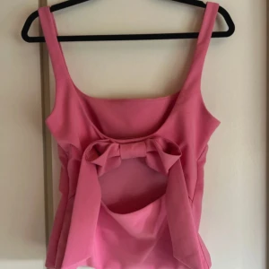 Rosa linnetopp med rosett och cut-out - Supersöt rosa linnetopp med breda axelband och en stor rosett framtill. Toppen har en snygg cut-out detalj under bysten som ger en unik look. Figurnära passform och slät yta, perfekt för dig som vill sticka ut lite extra. Den är helt ny och endast använd en gång där av priset! 