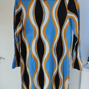 Retro mönstrad klänning i blått - Cool retroklänning med vågigt mönster i blått, svart, vitt och orange. Klänningen har trekvartsärm och är i en rak, normal passform. Perfekt för dig som gillar färg och unika prints. Materialet känns mjukt och lätt mot huden.