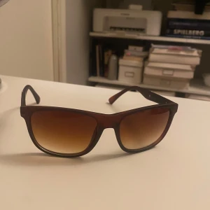 Bruna solglasögon Emporio Armani - Snygga solglasögon från Emporio Armani med brun plastbåge och rektangulär form. Glasen är tonade i brunt och bågarna har gulddetaljer samt logga på sidorna. Perfekt accessoar för att höja din stil och skydda ögonen mot solen.