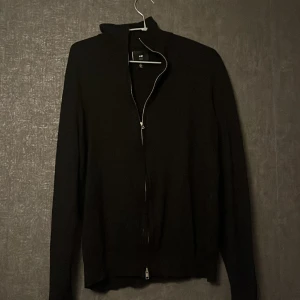 Svart slim kofta från H&M - Svart kofta från H&M i slim fit-modell med hel dragkedja framtill. Tröjan har lång ärm och en enkel, stilren design som passar perfekt till jeans eller chinos. Materialet är mjukt och skönt, troligtvis bomull eller polyester. Snygg att ha öppen eller stängd.