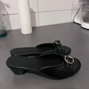 Svarta klackar / sandaler - Snygga svarta sandaler från Parda med låg klack och öppen tå. Skorna har silverfärgade spännen framtill med logga, vita kontrastsömmar och är tillverkade i skinn. Perfekta för dig som gillar stilrena och klassiska vibes med en touch av lyx. JÄTTESKÖN och mjuk sula. Super nice att gå i. HELT NYA!! Köpt för 599 på marknad 