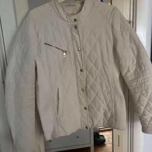 Vit quiltad jacka Holly & Whyte - Snygg vit quiltad jacka från Holly & Whyte med guldfärgade knappar och dragkedja. Jackan har långärmade ärmar, ribbade paneler på sidorna och en cool ståkrage. Perfekt för dig som gillar stilren och klassisk look med lite edge.