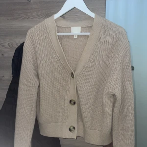 Beige stickad kofta med knappar - Säljer en beige stickad kofta från H&M med grov struktur och stora, snygga knappar framtill. Koftan har v-ringad hals och långa ärmar, perfekt för lager-på-lager. Passar dig som gillar chill och enkel stil.