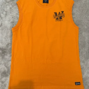 Orange linnetopp från Superdry - 🔥 Superdry Linnetopp 🔥 Cool orange linnetopp från Superdry 🧡 med trendigt tryck “Wildcats” framtill och ett stort svart 77 på ryggen ⚡. 	•	👕 Rund halsringning & ärmlös design för en sportig look 	•	🌞 Perfekt för sommaren, gymmet eller streetstyle 	•	🧵 Mjuk bomullskvalitet för hög komfort 	•	🏷️ Liten Superdry-label nedtill för den autentiska touchen  En färgstark topp som ger energi, attityd & stil – ett måste i garderoben! 