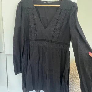 Svart kort klänning från Zara - Supersnygg svart klänning från Zara med v-ringning och lång ärm. Klänningen har broderade detaljer och spets längs bröstet och midjan, samt ett lätt prickigt mönster i tyget. Modellen är kort och har en luftig, lite volangig kjol. Perfekt för dig som gillar stilrena och feminina plagg. Säljs pga att den inte används