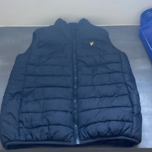 Svart dunväst från Lyle & Scott  - Svart dunväst från Lyle & Scott med gul broderad logga på bröstet. Västen har quiltad design, hög krage och dragkedja framtill. Perfekt för lager-på-lager och chill stil. Materialet är syntet och dun, vilket gör den både lätt och varm.